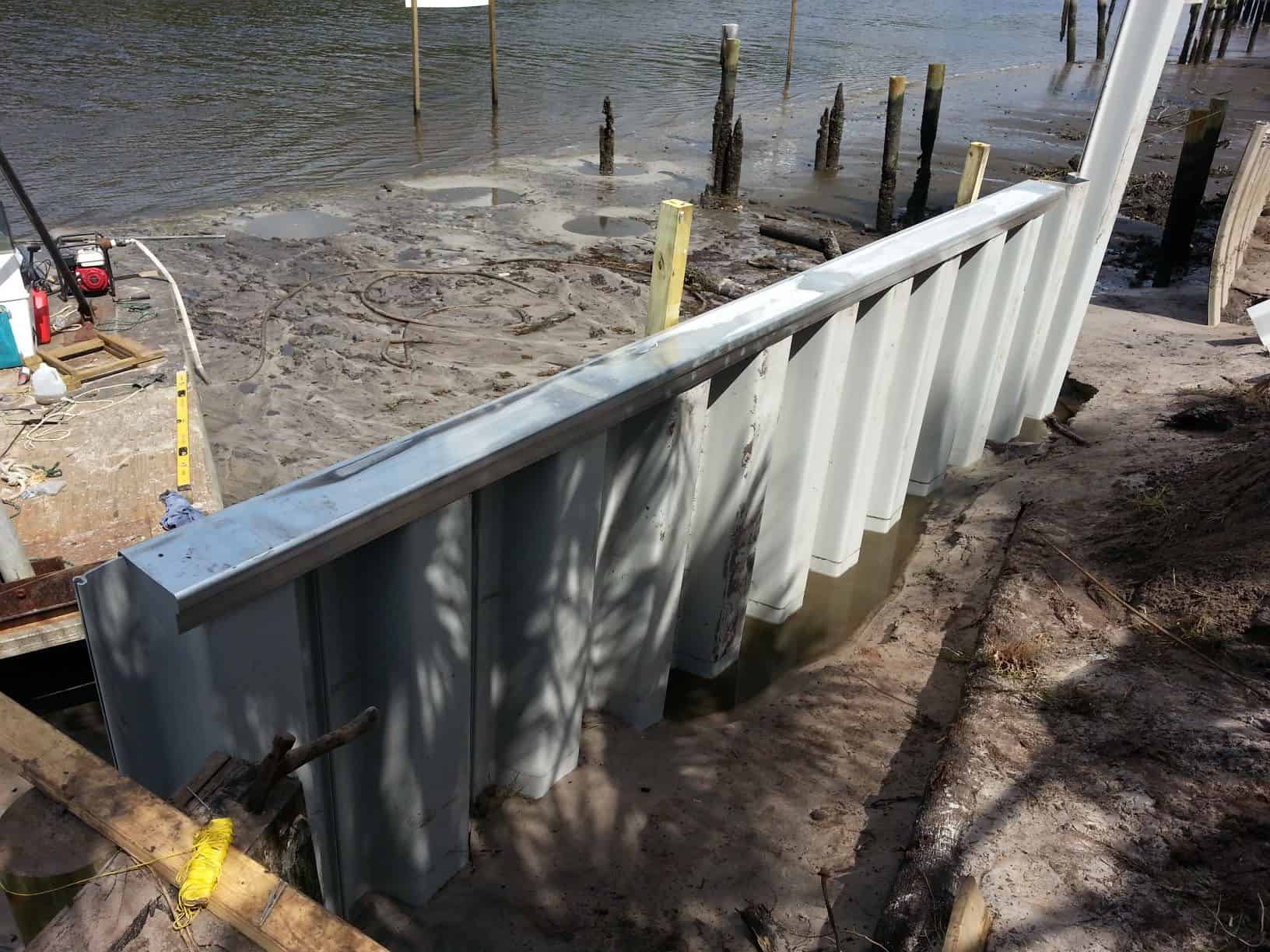 Newly driven vinyl sheet pile seawall with steel cap along a sandy shoreline, old pilings behind