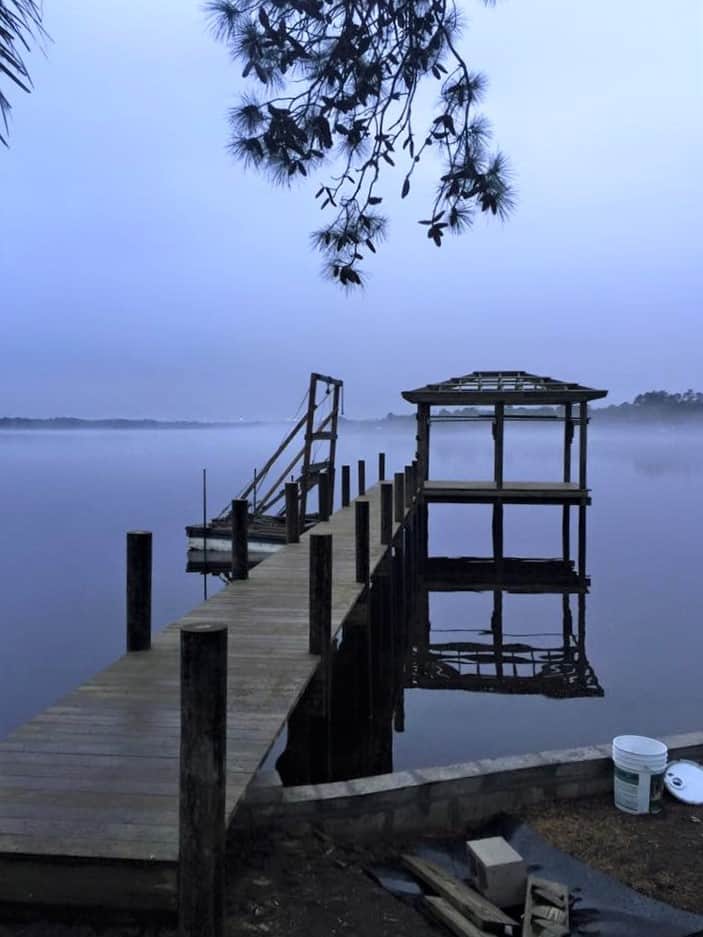 Dock walkway and open boathouse frame silhouetted over still water at dawn with ground fog