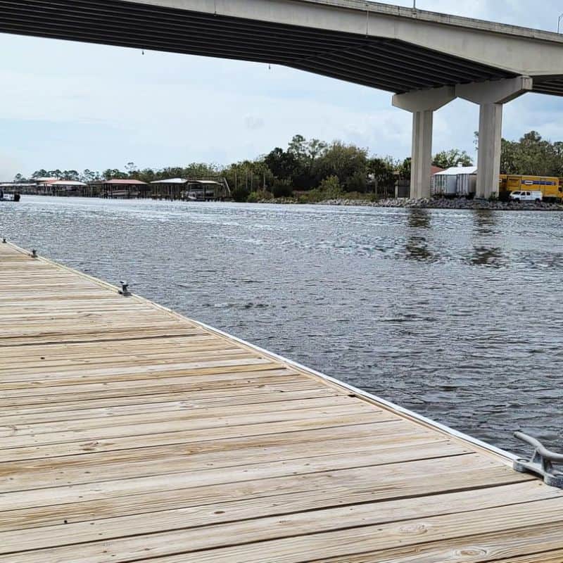New pressure-treated dock decking with mooring cleats beside a waterway under a concrete highway bridge