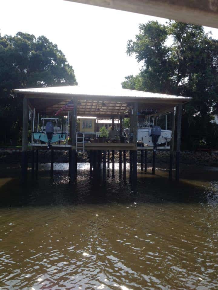 Covered boathouse on pilings with two outboard boats raised on lifts above tannic water