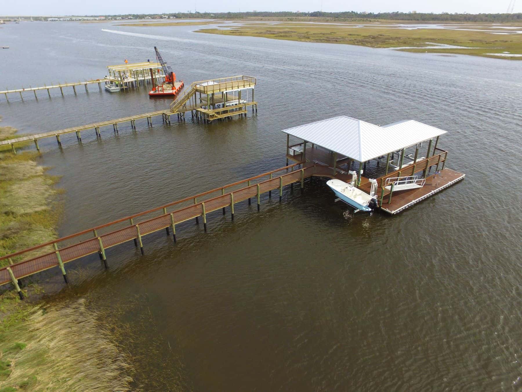 Aerial of a marsh-front boathouse and long pier, with a crane barge working on another dock
