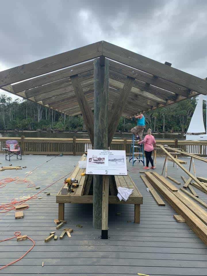 Workers building a timber pavilion roof on a gray composite dock deck amid scattered lumber