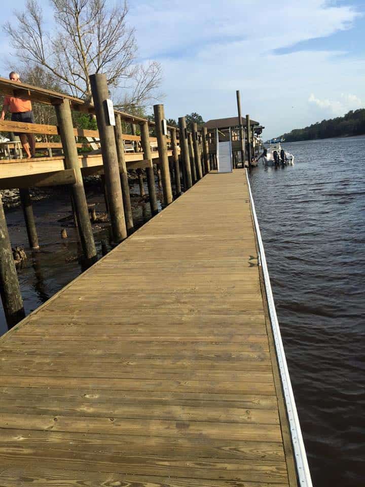 Long wood dock walkway running past pilings toward a covered structure and boat on the river