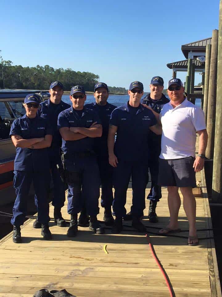 Group of uniformed Coast Guard personnel and a man in white posing on a wood dock