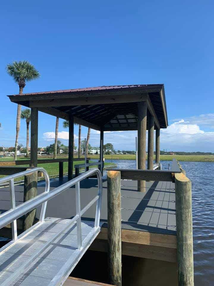 Covered dock with dark metal roof, aluminum gangway and railing over marsh water under blue sky