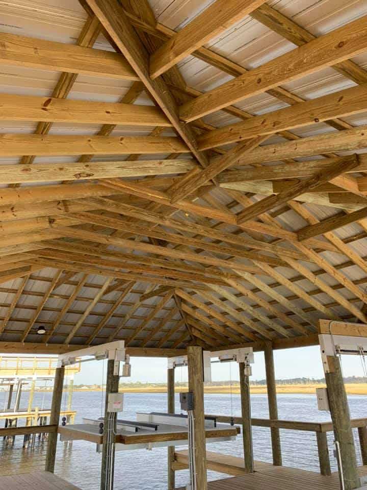 Looking up at a boathouse's exposed wood rafters and metal roof panels above empty lift bunks
