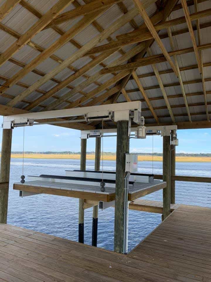 Empty four-post boat lift hanging on cables inside a wood-framed boathouse over a marsh