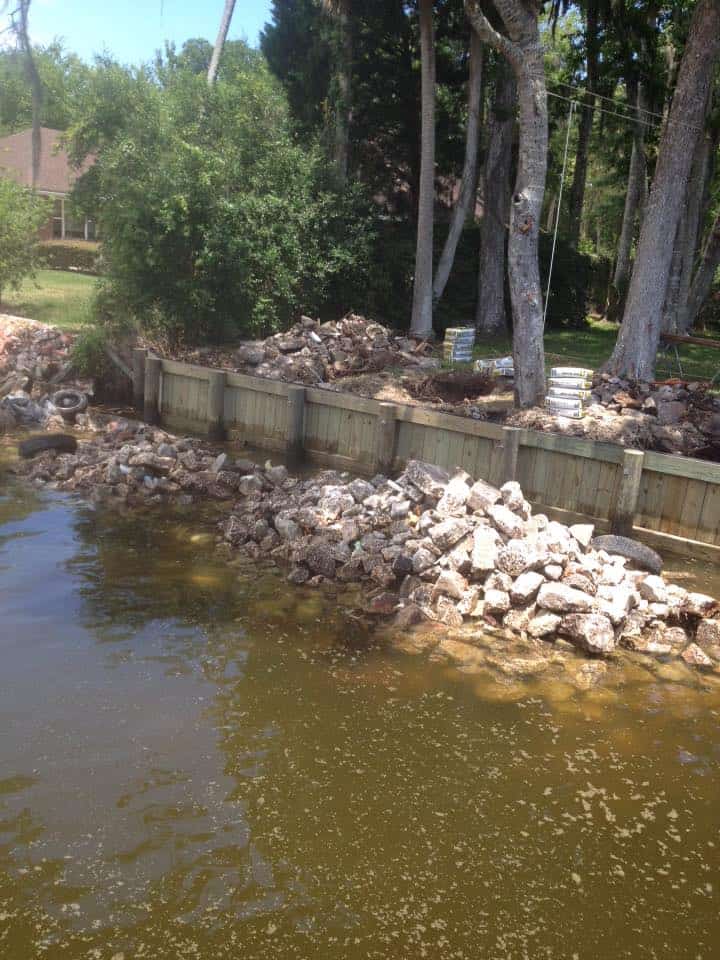 New wooden bulkhead behind a bank of broken concrete rubble along a shaded lakeshore