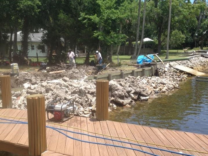 Two workers on shore behind a new bulkhead and concrete rubble, seen from a dock