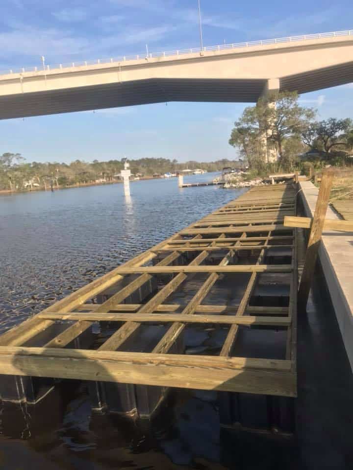 Dock joist framing on dark bulkhead sheeting beside a concrete wall, highway bridge overhead