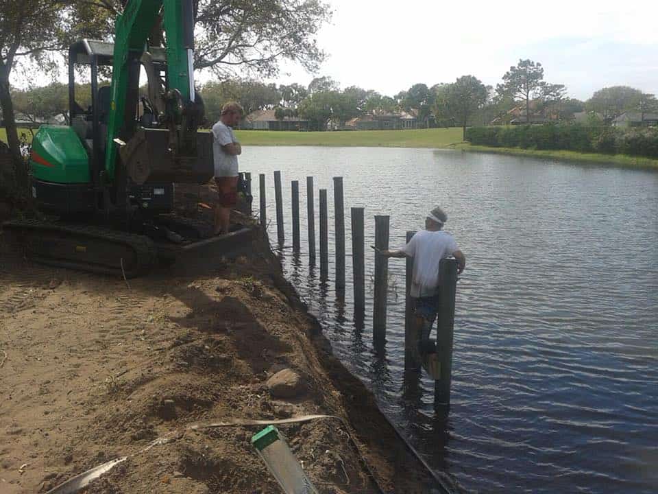 Two workers set pilings into a pond bank beside a mini excavator on shore