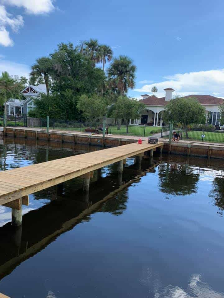 New wood walkway dock on pilings extending into a calm canal past a timber bulkhead and homes