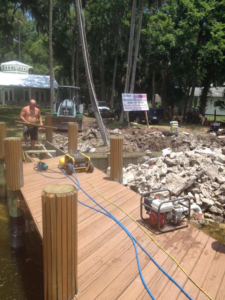 Worker on a new dock beside broken concrete rubble, contractor yard sign and excavator behind