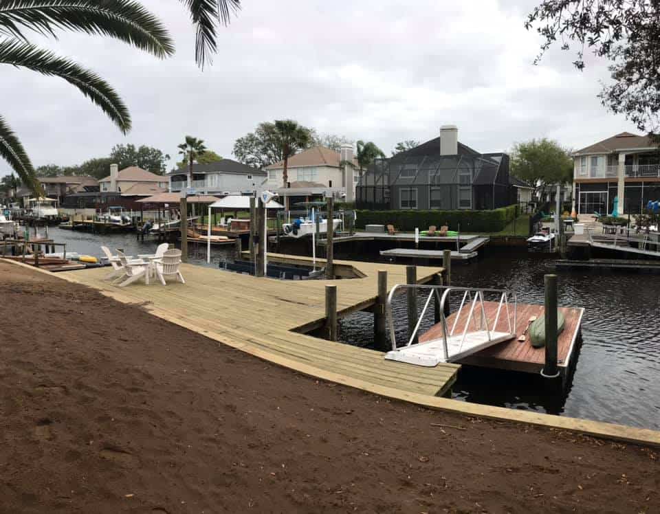 Wide wooden dock deck along a canal shoreline with lawn chairs and an aluminum gangway