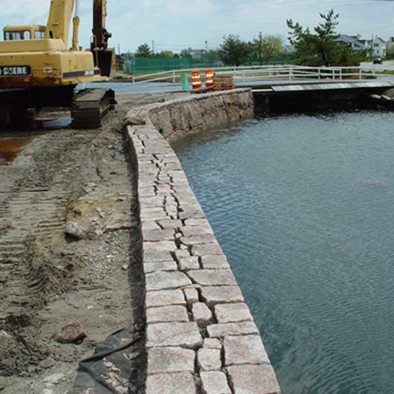 Stacked stone block seawall curving along a waterway with a John Deere excavator on the bank