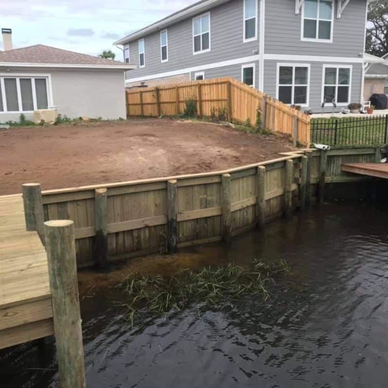 New timber seawall along a backyard canal with bare graded dirt behind and houses above