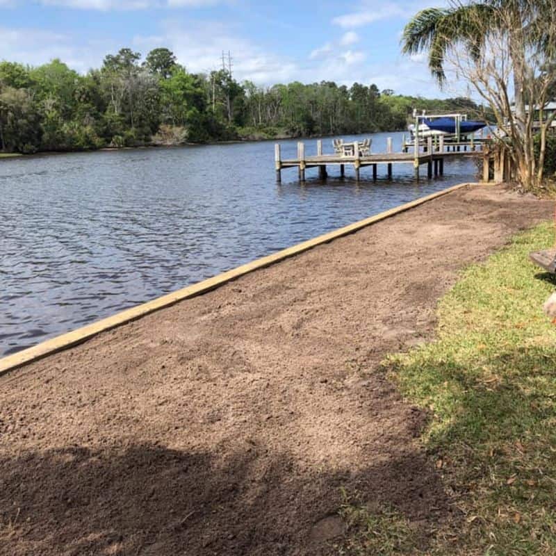 Low timber seawall edging a freshly graded dirt yard, wood dock and covered boat lift beyond