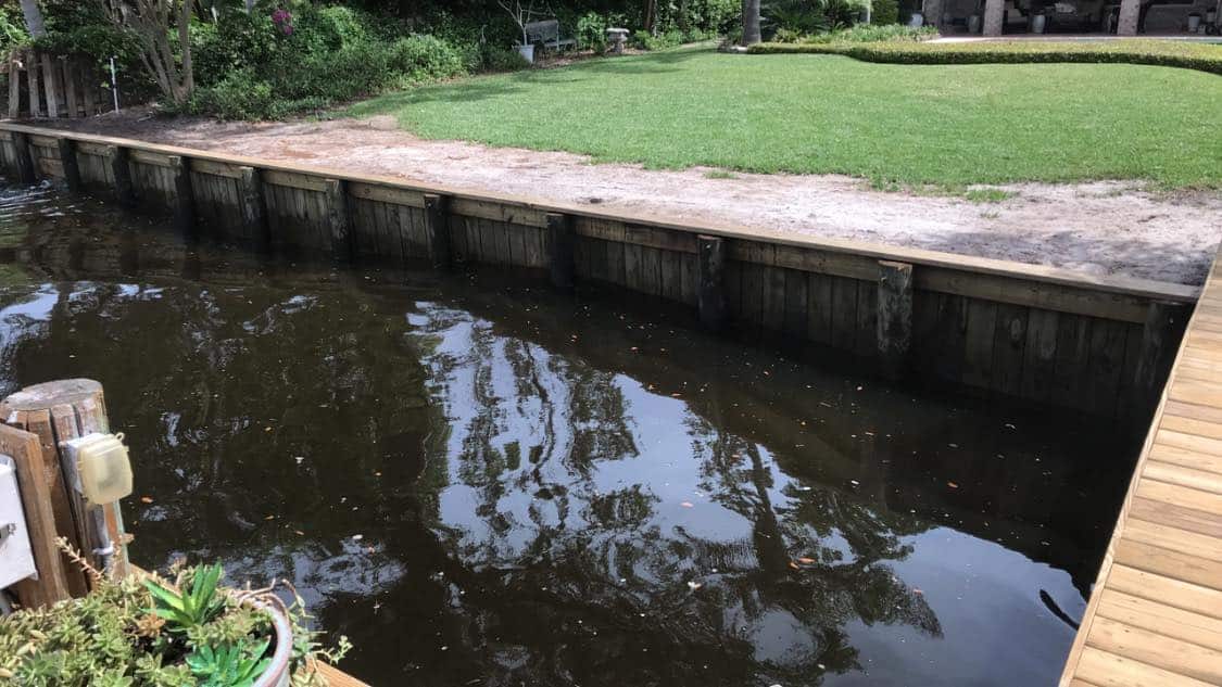 Long timber seawall with tie-back plates running beside a green lawn and shaded canal